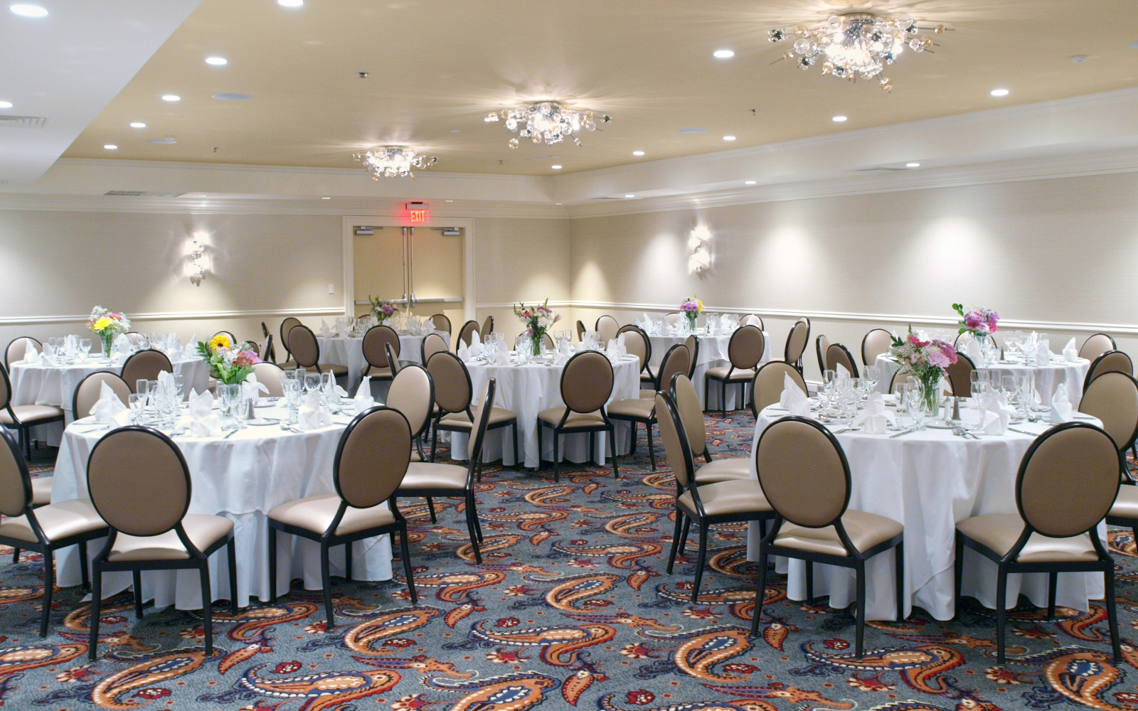 A large meeting room with round tables set for dining, adorned with floral arrangements and surrounded by beige chairs, under a ceiling with recessed lighting and chandeliers.