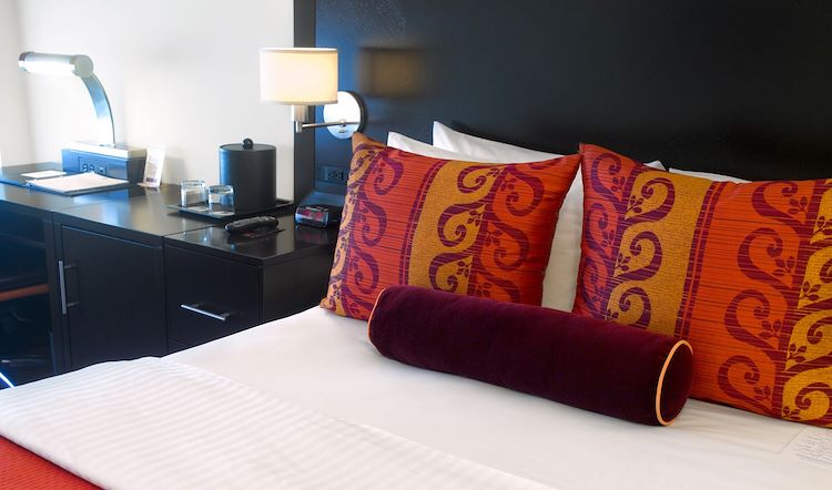 A hotel room bed with white sheets, orange and purple pillows, and a black desk with a lamp.